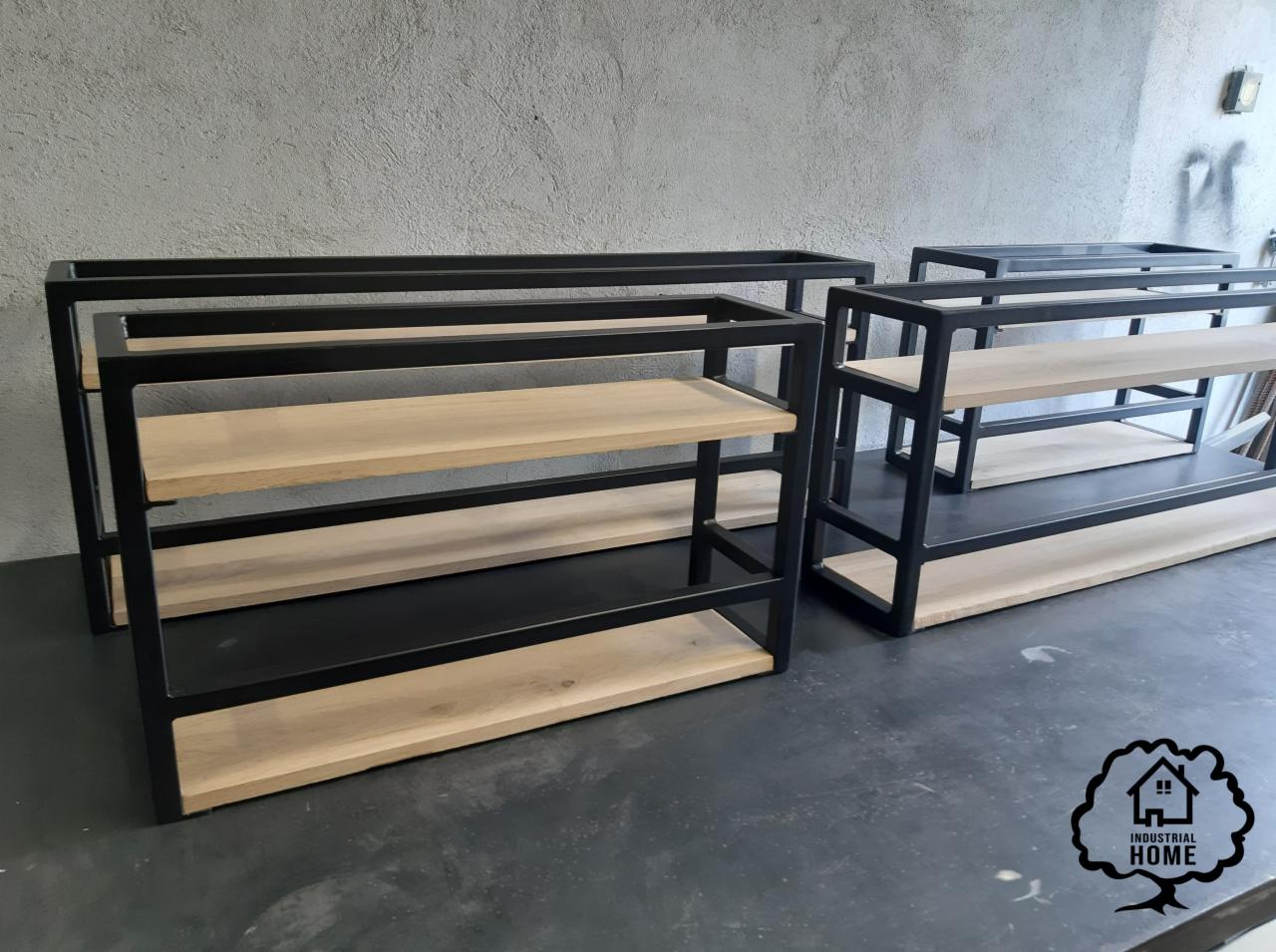 Metalowe regały magazynowe z drewnianymi półkami, czarne ramy, surowy beton w tle, logo producenta w prawym dolnym rogu.