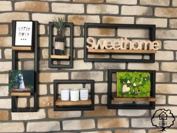 Zestaw dekoracyjnych, metalowych półek ściennych w stylu industrialnym z drewnianymi blatami, zamontowanych na ścianie z cegły, na których znajdują się ramki ze zdjęciami, wazon z rośliną, świeczki...