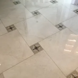 Jasne, marmurkowe płytki podłogowe ułożone ukośnie, z dekoracyjnymi kwadratami z motywem słońca w rogach, odbijające światło wpadające przez okno.