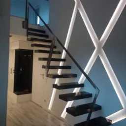 Nowoczesne schody wspornikowe z czarnymi stopniami i metalową balustradą, oświetlone dekoracyjnymi liniami LED w kształcie krzyży na ścianie.