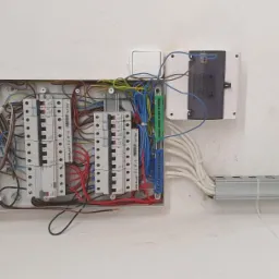 Otwarta skrzynka elektryczna z bezpiecznikami i przewodami na białej ścianie, widoczne szyny montażowe.