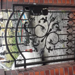 Czarna, metalowa furtka z ozdobnymi elementami liściastymi i geometrycznymi, zamontowana między dwoma ceglanymi słupkami. Widoczna skrzynka na listy. W tle zieleń i zaparkowany samochód.