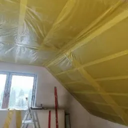 Poddasze w trakcie ocieplania, widoczna folia paroizolacyjna, drabina i poziomice przy ścianie, okno w tle.