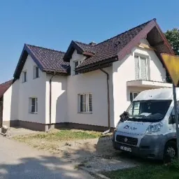 Wykończony dom jednorodzinny z jasną elewacją, brązowym dachem i okiennicami, zaparkowany biały samochód dostawczy z logo firmy w tle, słoneczny dzień.