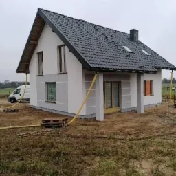 Dom jednorodzinny w trakcie prac elewacyjnych, widoczne fragmenty ocieplenia, szare pasy na ścianach, żółte węże i palety na ziemi, samochód dostawczy w tle.