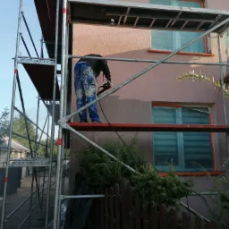 Pracownik na rusztowaniu szlifuje ścianę budynku przed malowaniem, okna zabezpieczone folią, widoczne plamy farby na spodniach pracownika.