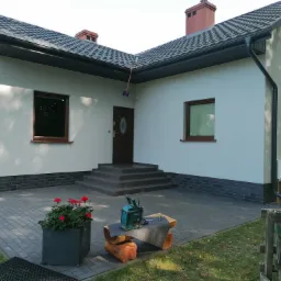 Nowo otynkowany dom jednorodzinny z szarą dachówką i ciemnymi oknami, widoczne schody wejściowe, dekoracyjny pas z płytek przy gruncie, na pierwszym planie kwiaty w donicy i radio budowlane.