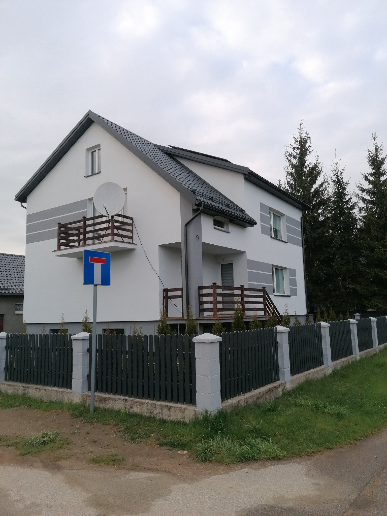 Dwukondygnacyjny dom z białymi ścianami i szarymi akcentami, balkonem z drewnianą balustradą i anteną satelitarną, otoczony szarym ogrodzeniem z betonowymi słupkami, przy którym stoi znak drogowy...