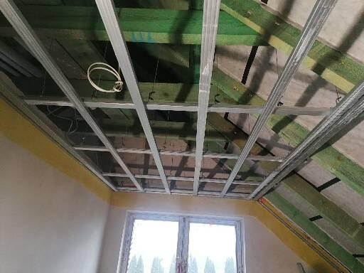 Metalowa konstrukcja sufitu podwieszanego z widocznymi elementami drewnianymi poddasza, okno w tle.