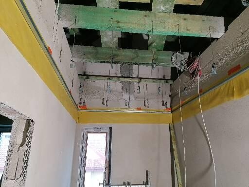 Wnętrze w trakcie prac wykończeniowych: widoczne belki stropowe, instalacja elektryczna i przygotowane ściany z żółtą taśmą zabezpieczającą, okno w tle.