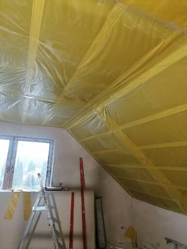 Poddasze w trakcie ocieplania, widoczna folia paroizolacyjna, drabina i poziomice przy ścianie, okno w tle.