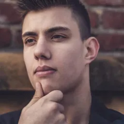 Portret młodego mężczyzny z ręką pod brodą, zamyślony wyraz twarzy, ceglana ściana w tle.