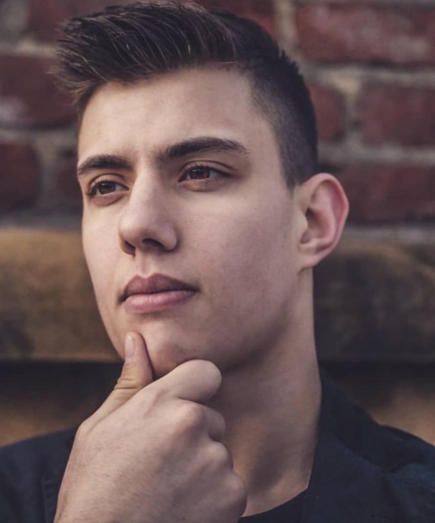 Portret młodego mężczyzny z ręką pod brodą, zamyślony wyraz twarzy, ceglana ściana w tle.