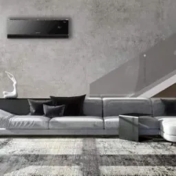 Czarny klimatyzator ścienny w nowoczesnym salonie z szarą sofą narożną, betonową ścianą i schodami z szklaną balustradą.