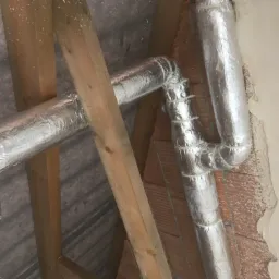 Fragment instalacji rekuperacji w przestrzeni poddasza z widocznymi przewodami wentylacyjnymi owiniętymi folią aluminiową i przymocowanymi plastikowymi opaskami zaciskowymi, drewnianą konstrukcją...