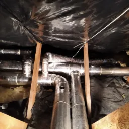 Izolowane, srebrne rury wentylacyjne w przestrzeni poddasza, pod czarną folią, wsparte drewnianymi belkami.