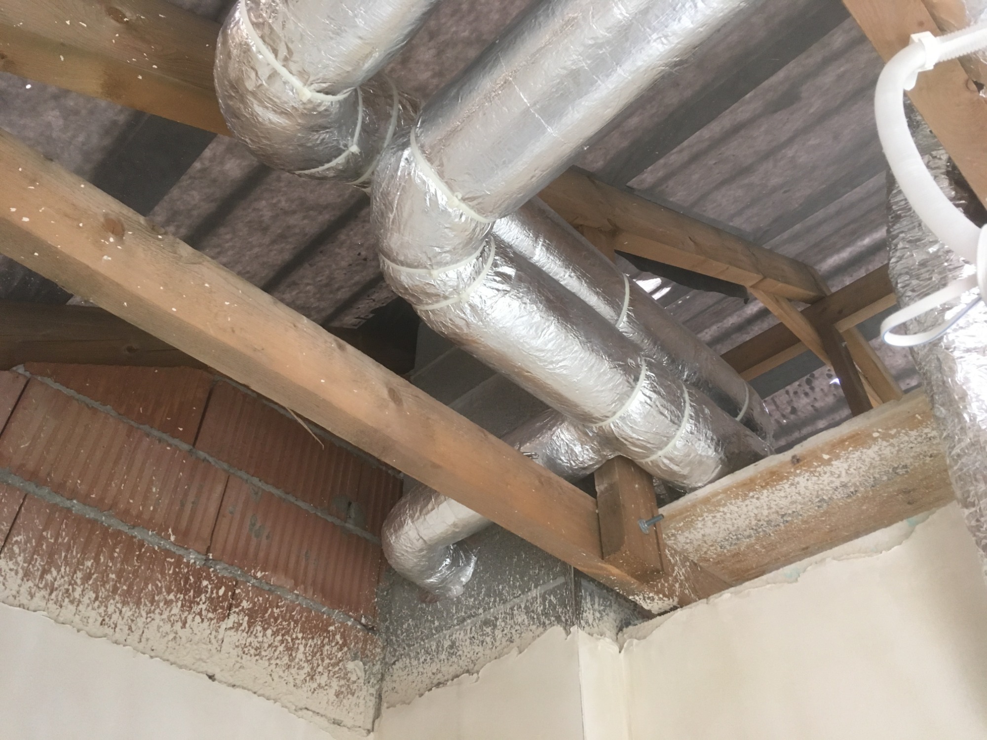 Srebrne, izolowane przewody wentylacyjne zamontowane pod stropem, widoczne drewniane belki konstrukcyjne i fragment ściany z cegły.