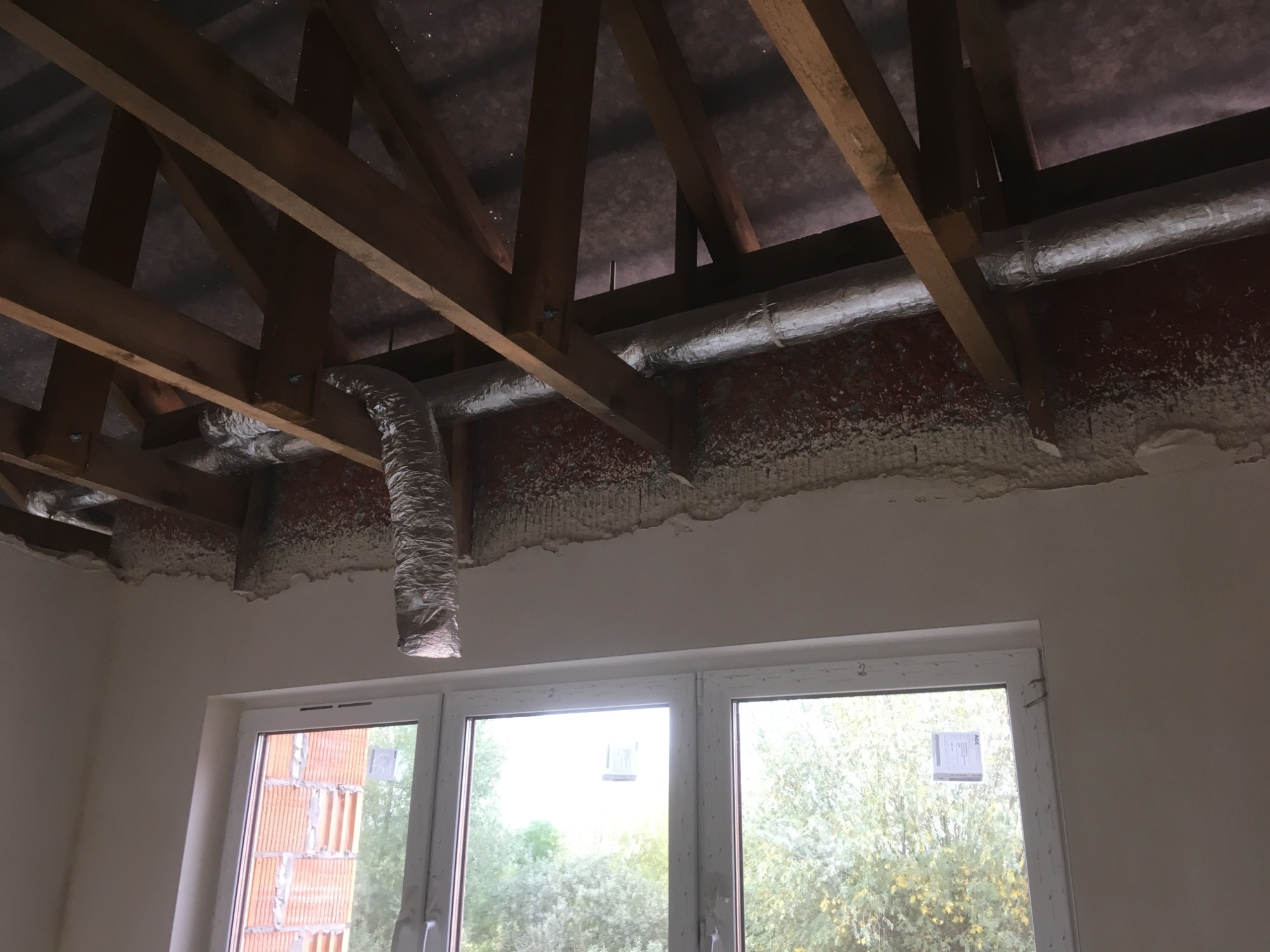 Montaż kanału wentylacyjnego z folii aluminiowej w nowym budynku, widoczna konstrukcja dachu i ocieplenie pianą, okno z widokiem na drzewa.