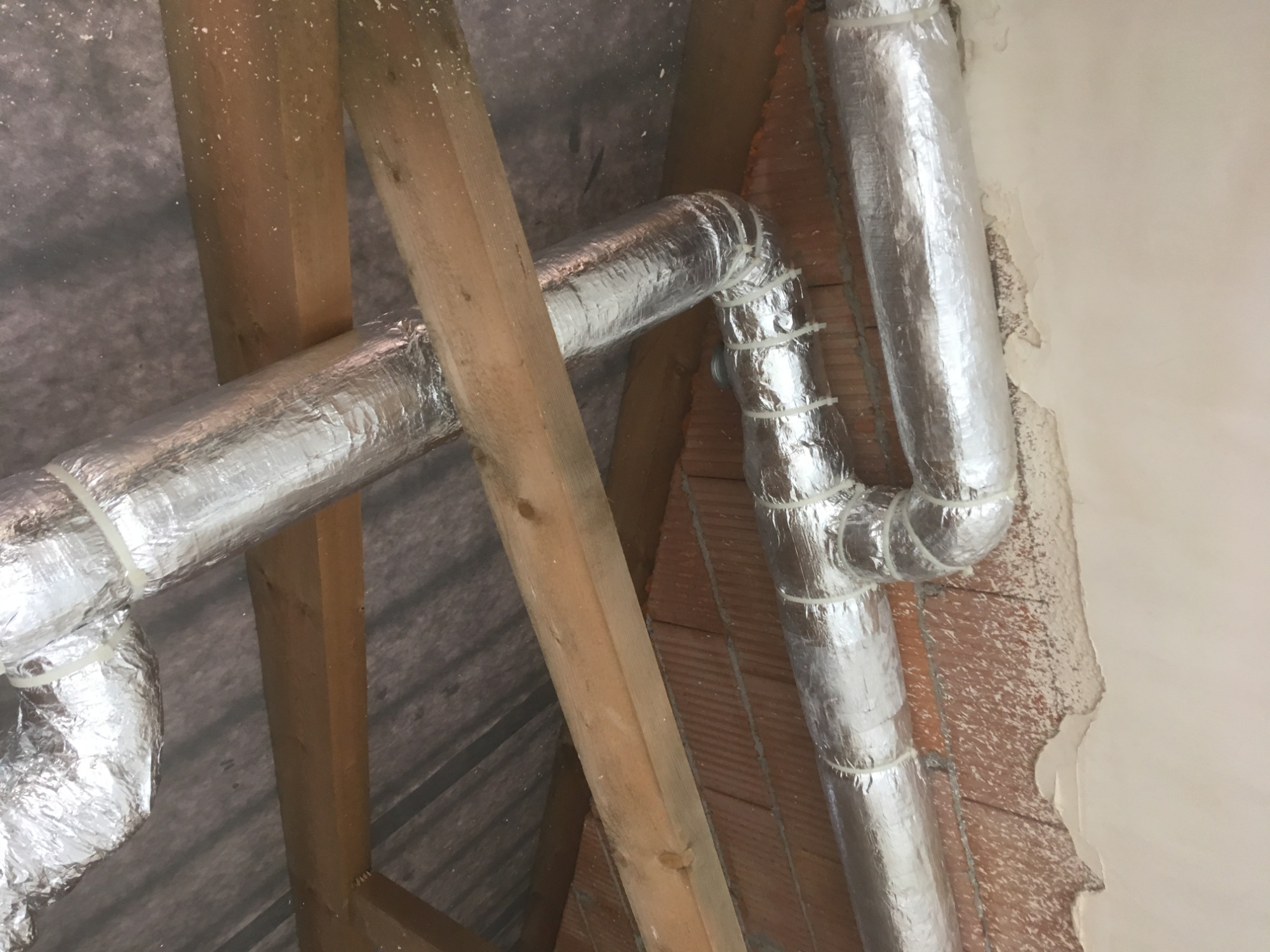 Fragment instalacji rekuperacji w przestrzeni poddasza z widocznymi przewodami wentylacyjnymi owiniętymi folią aluminiową i przymocowanymi plastikowymi opaskami zaciskowymi, drewnianą konstrukcją...