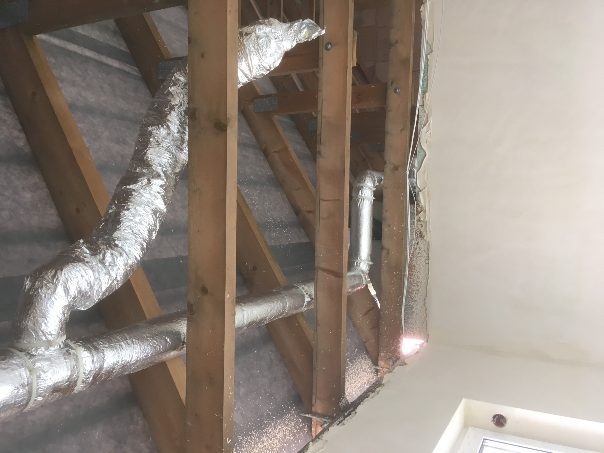 Montaż elastycznych, izolowanych przewodów powietrznych w przestrzeni poddasza, widoczne drewniane krokwie i fragment ściany po remoncie.