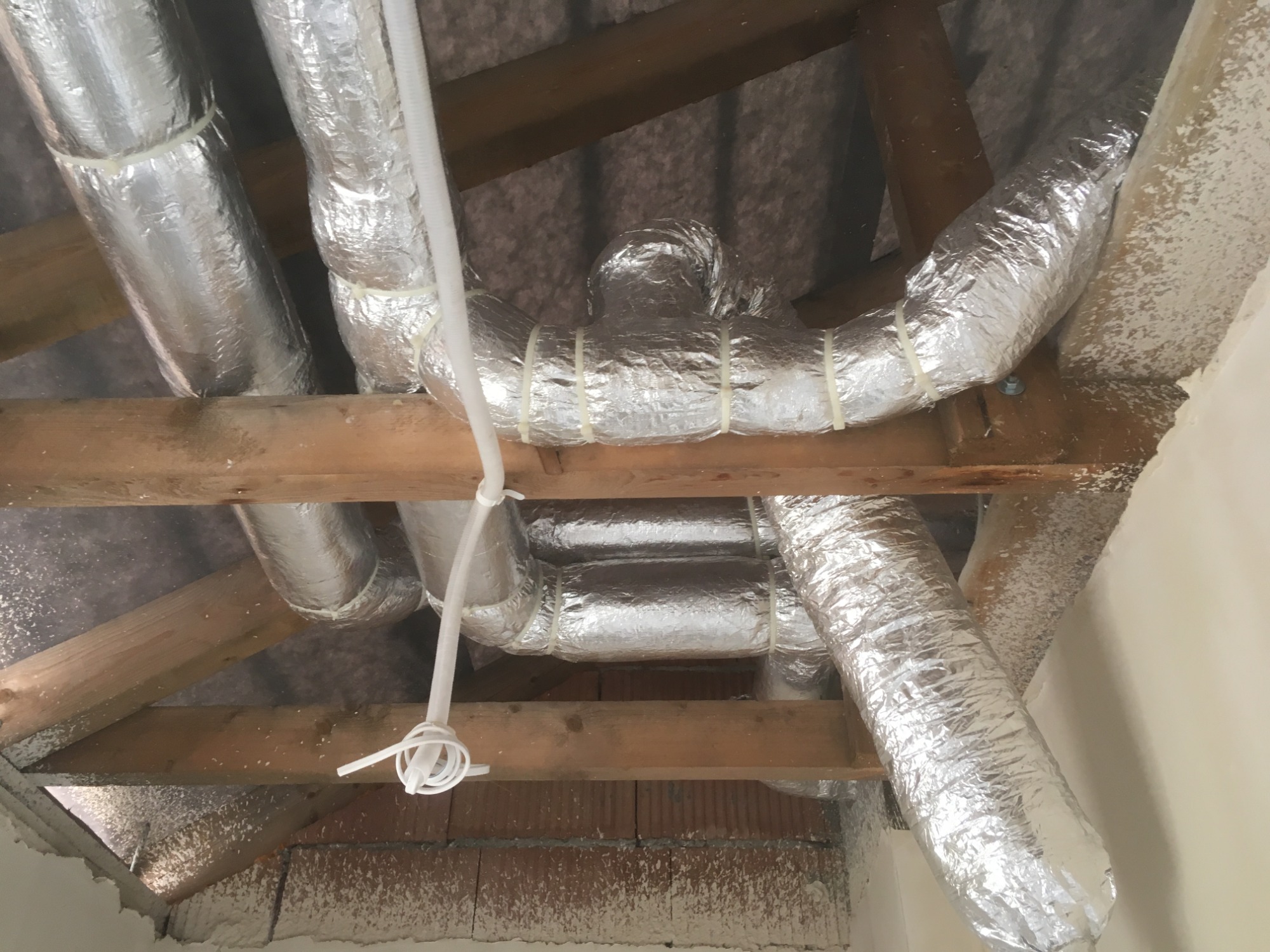 Srebrne, izolowane przewody wentylacyjne zamontowane pod drewnianą konstrukcją dachu, zabezpieczone białymi opaskami zaciskowymi.