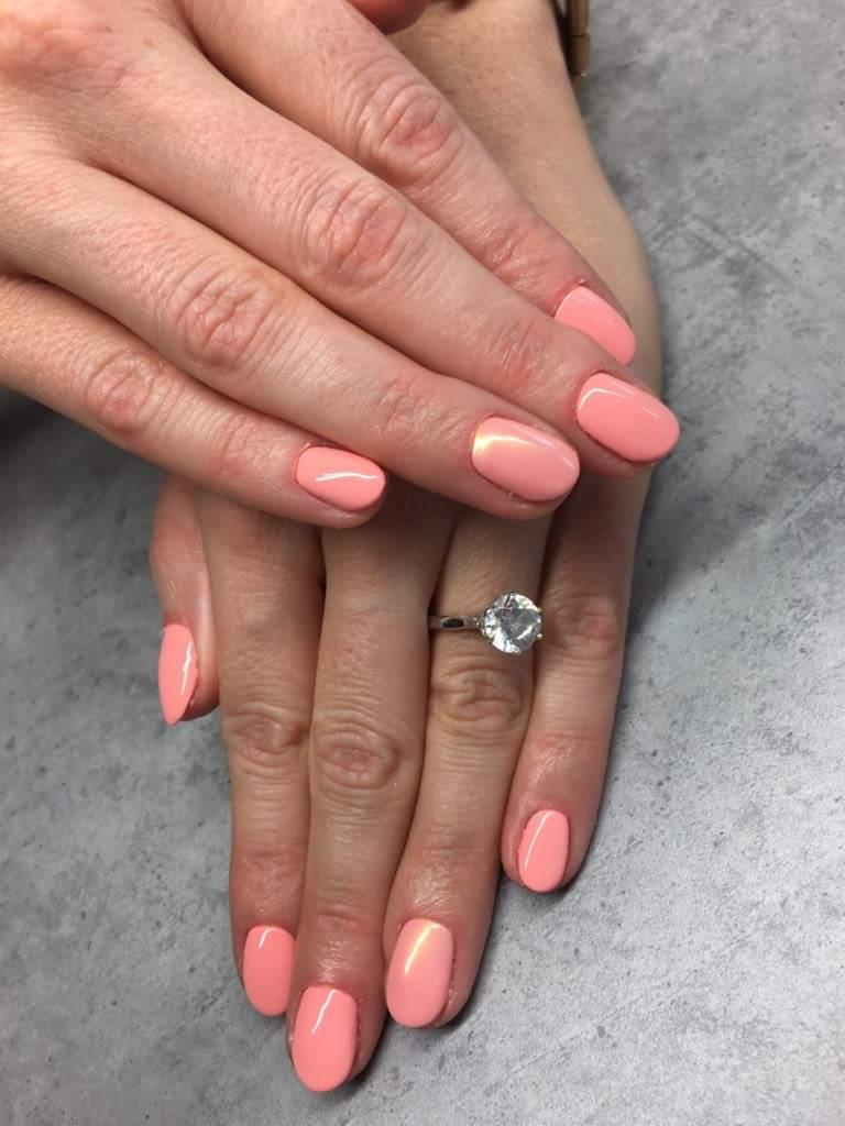 Dłonie z świeżo pomalowanymi paznokciami w kolorze koralowym z subtelnym połyskiem, pierścionek zaręczynowy z dużym brylantem na serdecznym palcu.