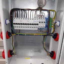 Wnętrze metalowej skrzynki rozdzielczej z zamontowanymi bezpiecznikami EATON, przewodami w różnych kolorach, złączami w kolorze czerwonym i niebieskim oraz kluczem do otwierania skrzynki na dnie.