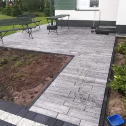 Ułożona kostka brukowa w odcieniach szarości tworząca ścieżkę i taras przy budynku z zielonymi ławkami i stołem, obrzeża z ciemnej kostki, rabata ziemi po lewej stronie.
