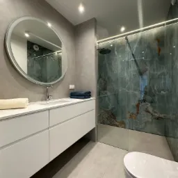 Nowoczesna łazienka w odcieniach szarości z akcentem marmurowej ściany prysznicowej, okrągłe lustro z podświetleniem LED nad białą szafką z umywalką.