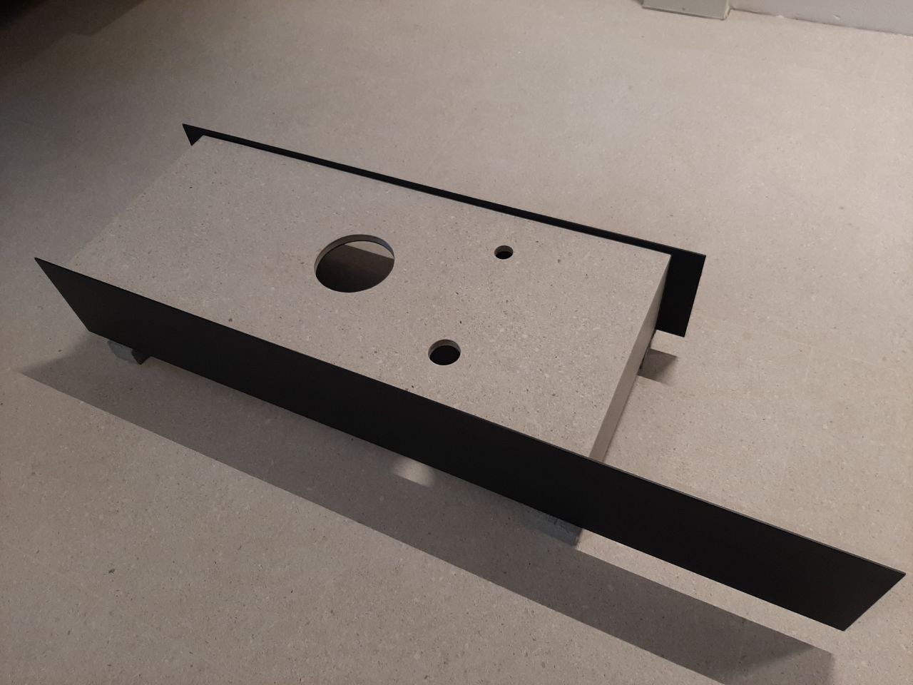 Blat z otworami, obudowany czarnymi listwami, leżący na podłodze. Widoczne dwa małe otwory i jeden większy, centralny.