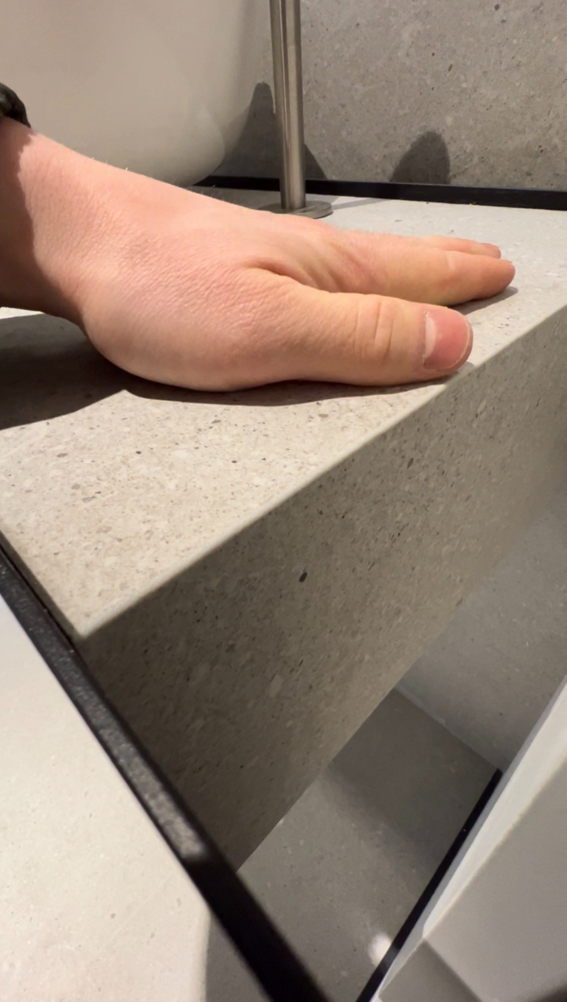 Dłoń spoczywająca na blacie z konglomeratu kwarcowego w łazience, widoczny fragment umywalki i stalowy słupek mocujący.