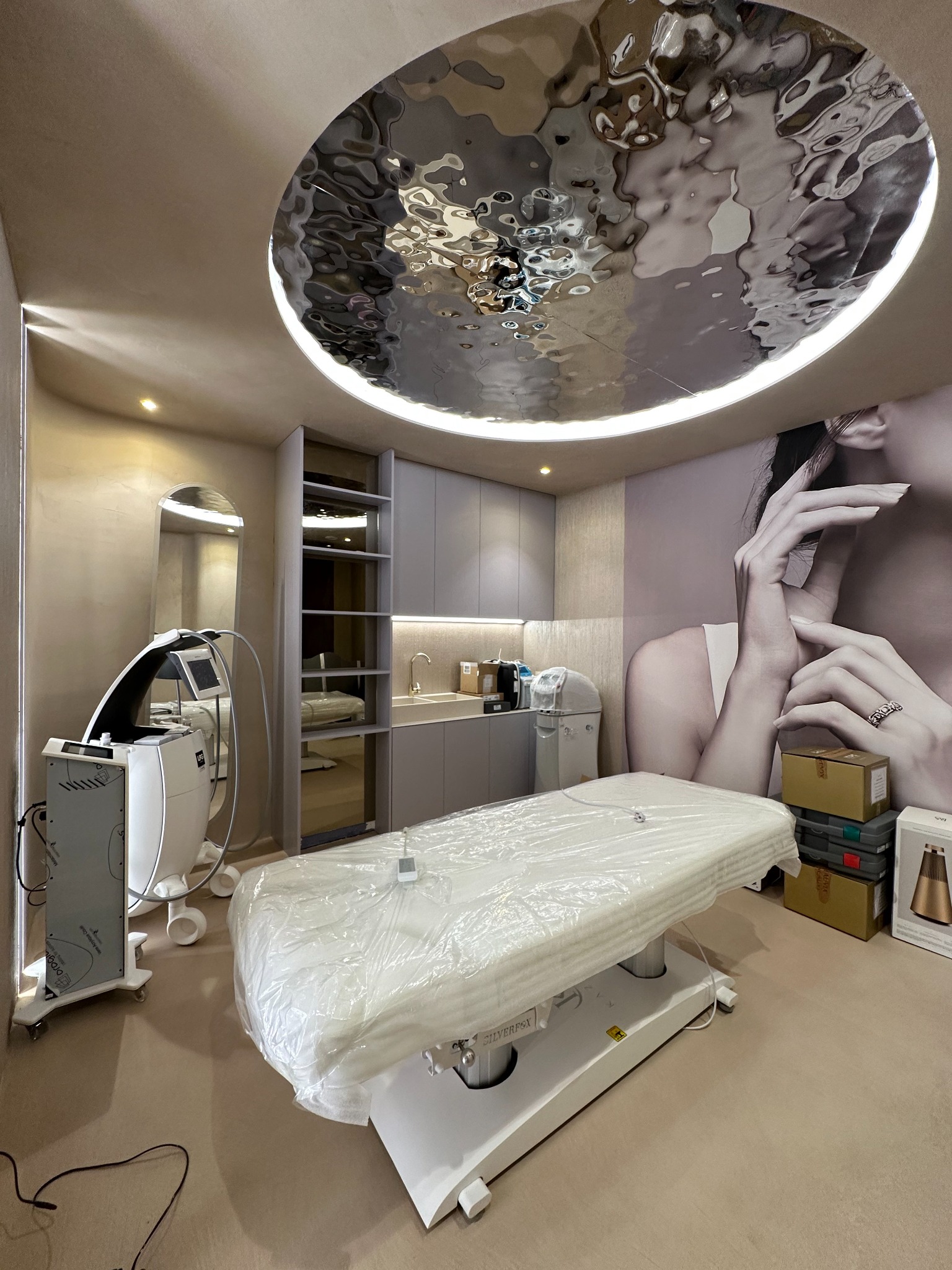 Kremowe wnętrze gabinetu kosmetycznego z nowoczesnym oświetleniem sufitowym, białym fotelem do zabiegów zabezpieczonym folią, sprzętem kosmetycznym i dekoracyjną tapetą z wizerunkiem kobiety.