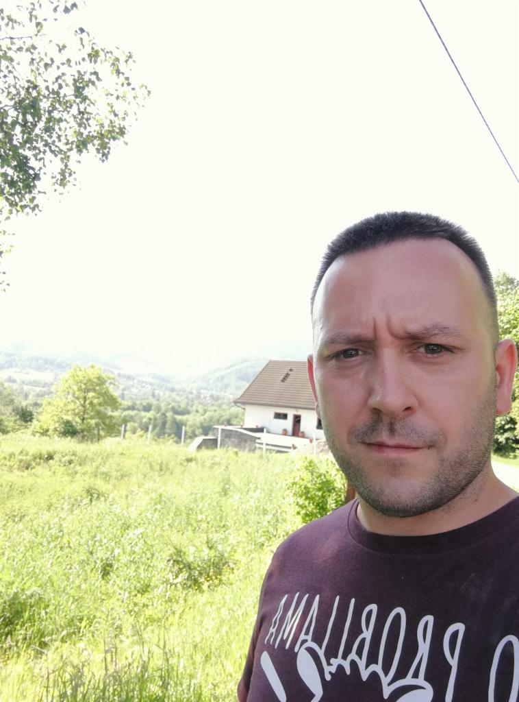 Mężczyzna na tle domu z szarą dachówką i zieleni. Ujęcie z dołu, widoczne wzgórza w oddali. Twarz mężczyzny wyraźna.