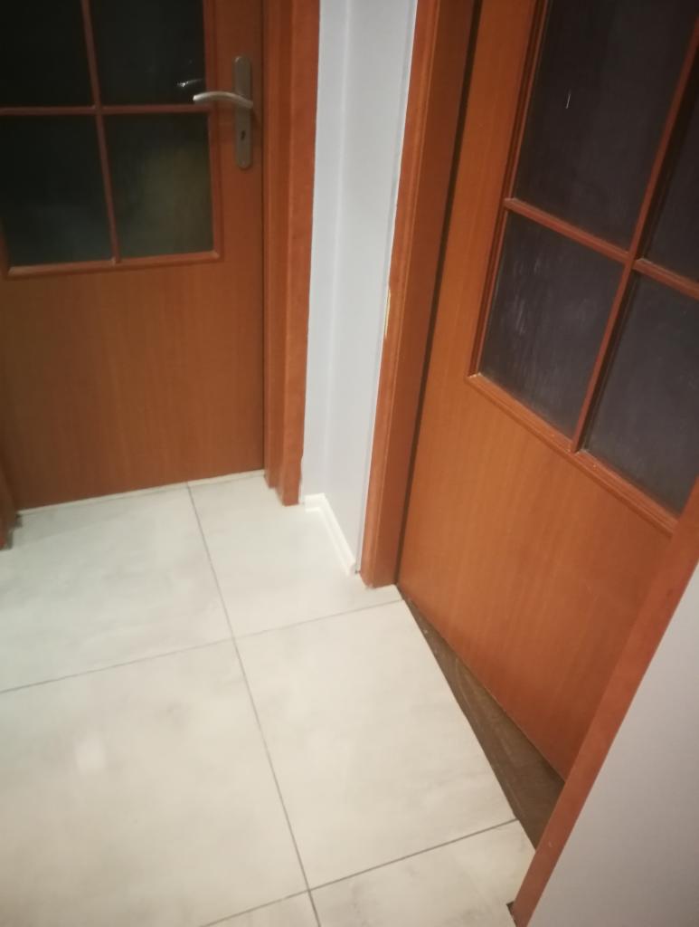 Dwie drewniane drzwi z szybami, widoczne pod kątem, na tle jasnych płytek podłogowych i szarej ściany.