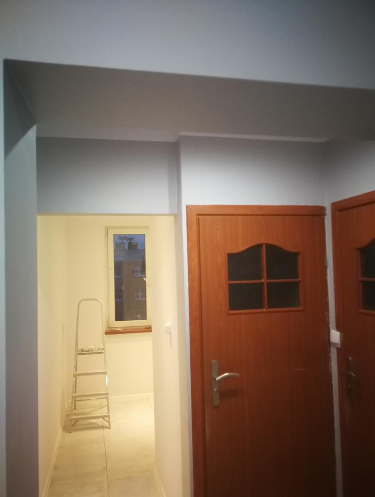 Korytarz w trakcie malowania na biało, widoczna drabina aluminiowa, pomalowane ściany i sufit, brązowe drzwi z szybkami.