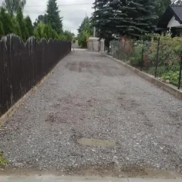 Nawierzchnia z szarego kruszywa na posesji, widoczna kałuża, obrzeża z kostki, w tle ogrodzenie z siatki i drewniany płot.