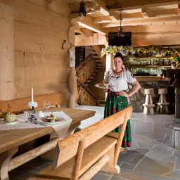 Wnętrze góralskiej restauracji: kelnerka w stroju ludowym z talerzem dań, drewniane meble, kamienny kominek, bar z alkoholem w tle.