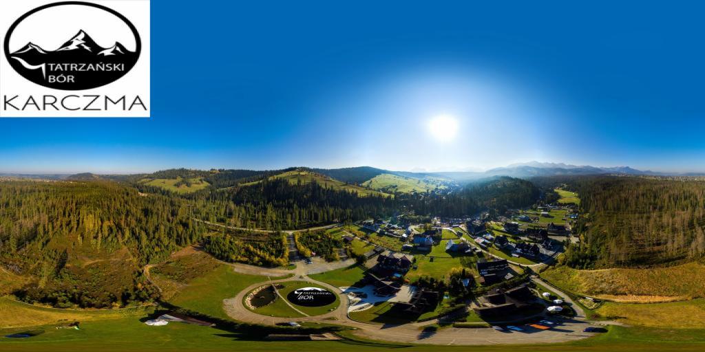 Panoramiczny widok z lotu ptaka na Tatrzański Bór Karczma, otoczony zielonymi wzgórzami, lasami i błękitnym niebem z jasnym słońcem.