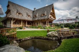 Drewniany budynek w stylu zakopiańskim z charakterystycznym dachem i oknami w kształcie oczu, otoczony zielonym trawnikiem, oczkiem wodnym z kamieniami i drewnianym płotkiem, częściowo widoczny...