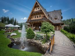 Drewniany budynek w stylu góralskim z fontanną na pierwszym planie, otoczony zielenią i drzewami, z widocznymi parasolami i stolikami w tle.