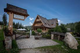 Drewniany budynek w stylu zakopiańskim z bramą wejściową i drogowskazem z drewnianym daszkiem, otoczony zielenią i kamiennymi elementami.