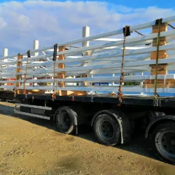 Transport elementów stalowych na paletach, zabezpieczonych łańcuchami, na naczepie ciężarówki, w słoneczny dzień.