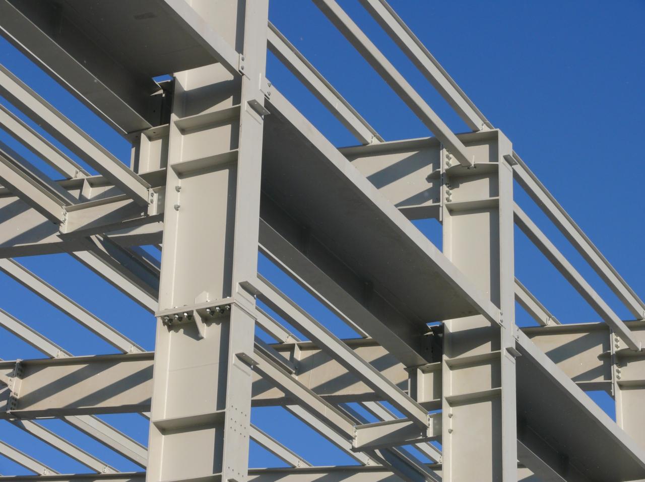 Szare, stalowe elementy konstrukcyjne hali w trakcie montażu, połączone śrubami na tle błękitnego nieba.
