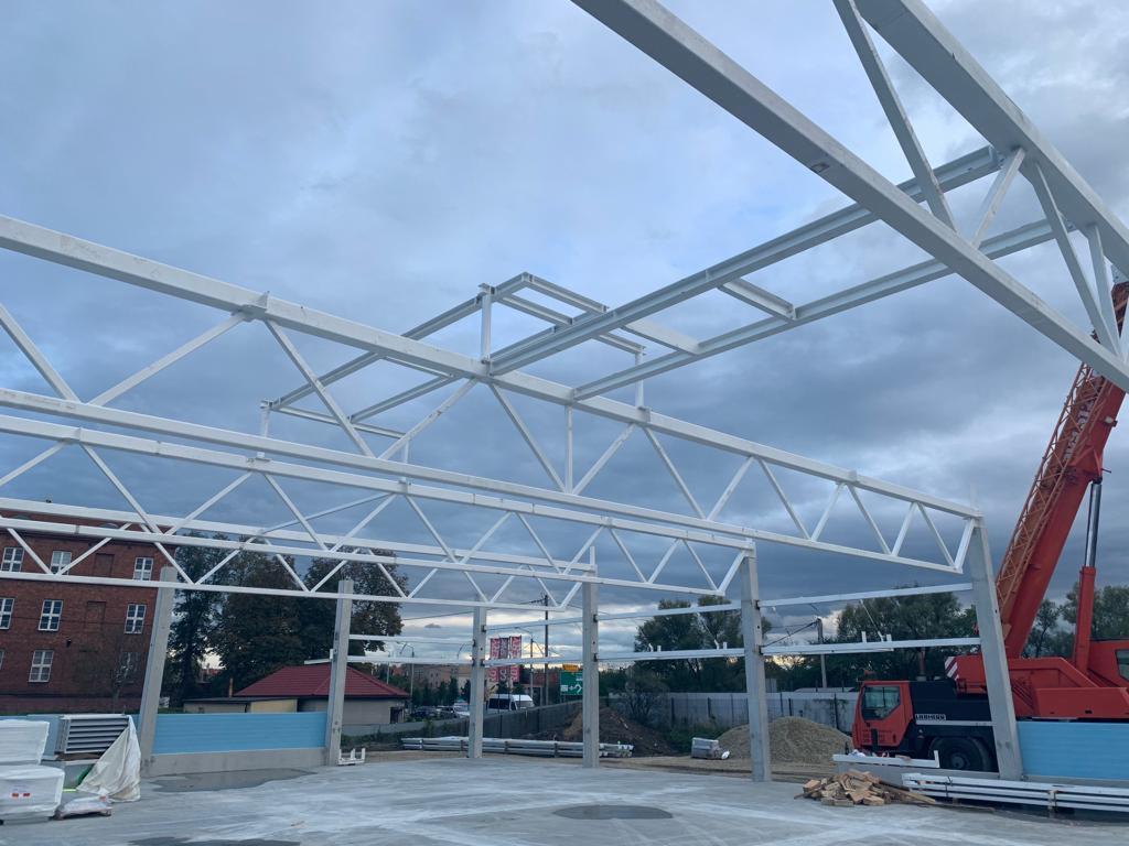 Konstrukcja stalowa hali w trakcie budowy, widoczny biały szkielet i dźwig Liebherr w tle, betonowa podstawa, pochmurne niebo.