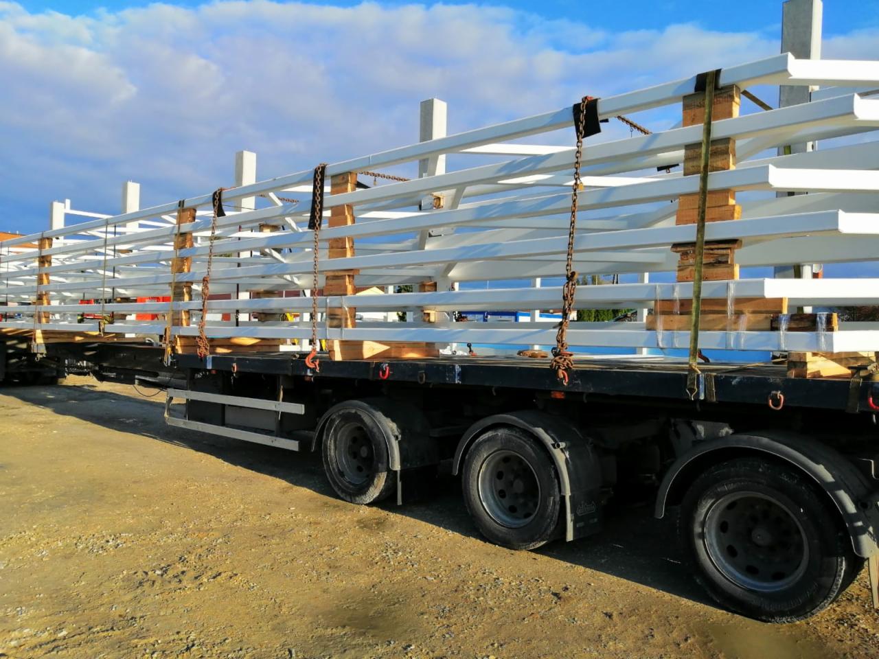 Transport elementów stalowych na paletach, zabezpieczonych łańcuchami, na naczepie ciężarówki, w słoneczny dzień.