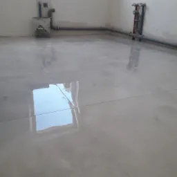 Świeżo położone, jasnoszare płytki podłogowe w surowym wnętrzu. Widoczne rury instalacji wodnej przy ścianach i wiadro po zaprawie budowlanej.