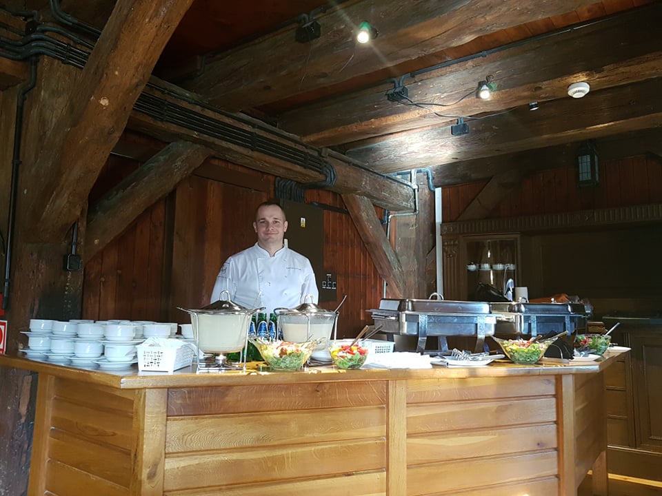 Szef kuchni w białym uniformie za drewnianym stołem z bufetem: białe filiżanki, srebrne naczynia z zupą krem i sałatki w szklanych misach, rustykalne wnętrze z drewnianymi belkami stropowymi.