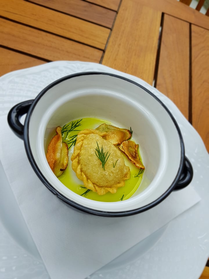 Pojedynczy pieróg z farszem, udekorowany koperkiem i chipsami warzywnymi, podany w białej miseczce z czarnymi uchwytami, w klarownym sosie na drewnianym stole.