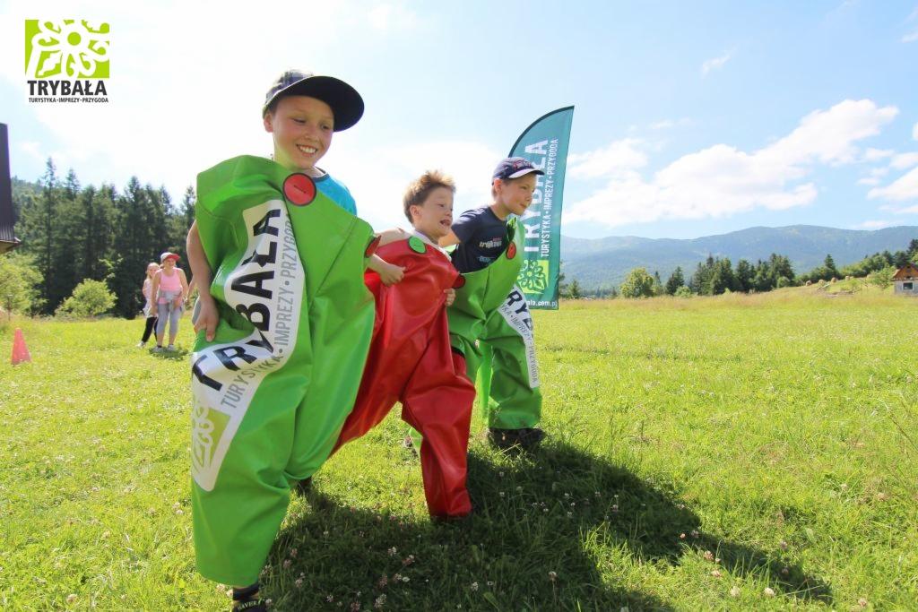 Dzieci w kolorowych workach z logo firmy Trybała Turystyka-Imprezy-Przygoda biorą udział w wyścigu na zielonej łące z widokiem na góry.