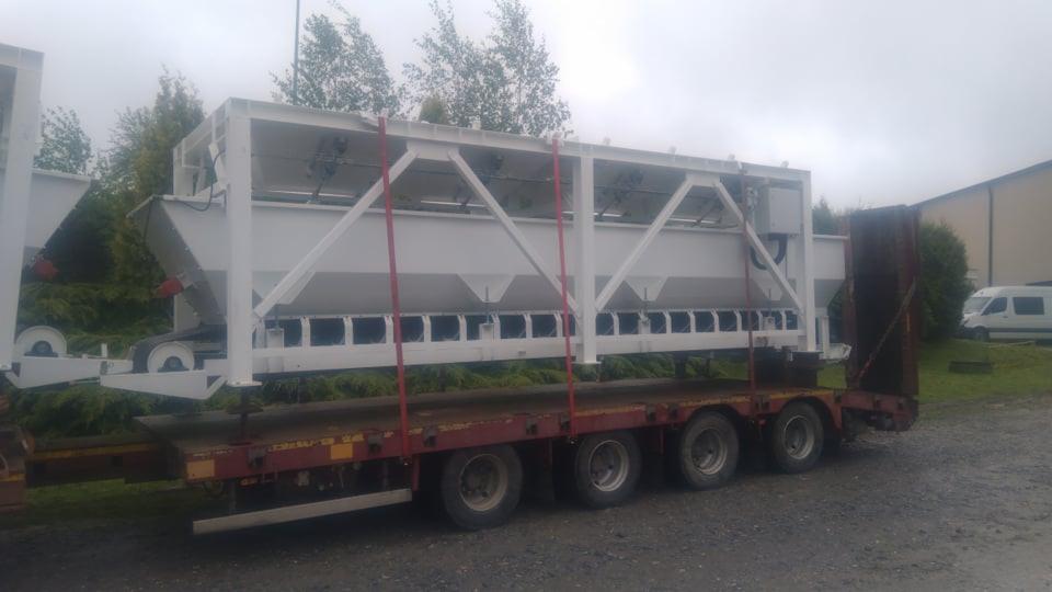 Biała konstrukcja stalowa z lejkowatymi zbiornikami, zabezpieczona czerwonymi pasami, transportowana na niskopodwoziowej przyczepie. W tle widoczne budynki i zieleń.